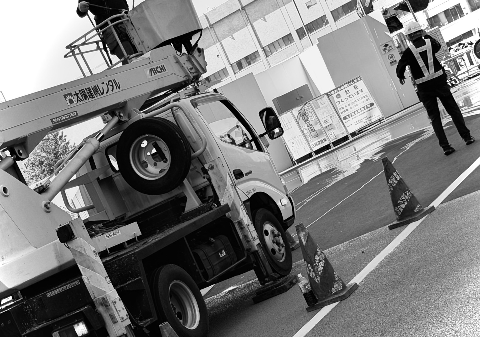 高所作業車がバケットを上げて作業する横で、コーンを置いた車線の車両を誘導する警備員