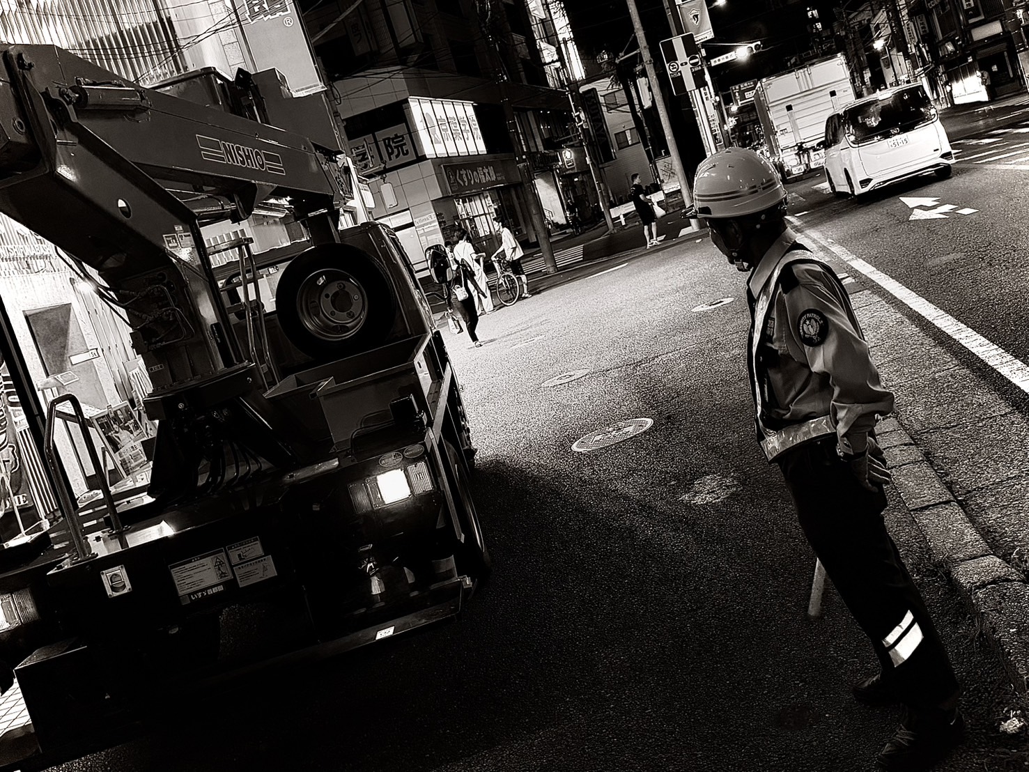 夜の市街地でクレーン車の作業中、路肩から歩行者と車両の動きを見守る警備員