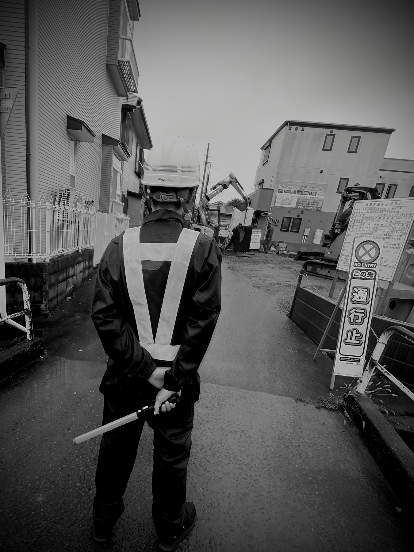 「通行止」の看板が立つ工事現場の入口で、誘導灯を手に立つ警備員の後ろ姿